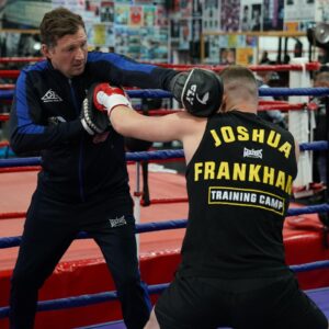 A boxing coach holds up pads while a boxer wearing a Joshua Frankham Training Camp vest throws a punch in a gym with posters on the walls and red, blue, and white ropes around the ring.