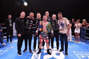 A male boxer wearing championship belts stands in a boxing ring, surrounded by his team and supporters. Some people are applauding in the background, and everyone is smiling for the photo.
